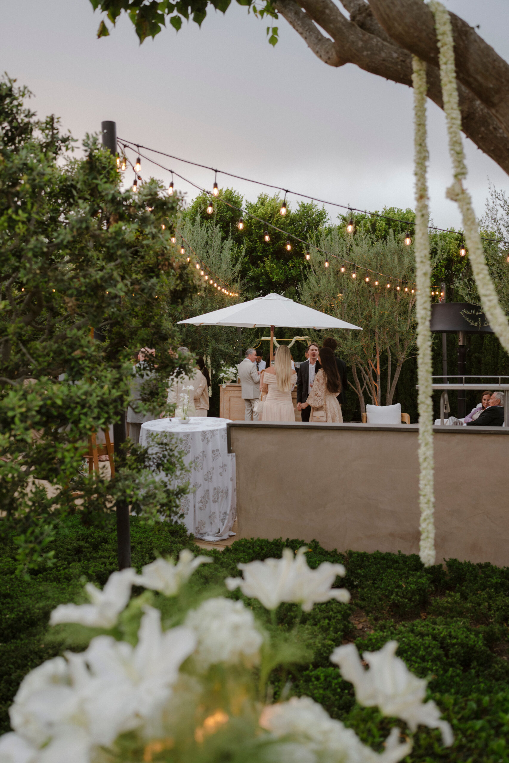 Outdoor wedding welcome party under string lights with guests mingling near the bar and floral decor, showcasing cohesive flower design ideas for a multi-day wedding weekend celebration.